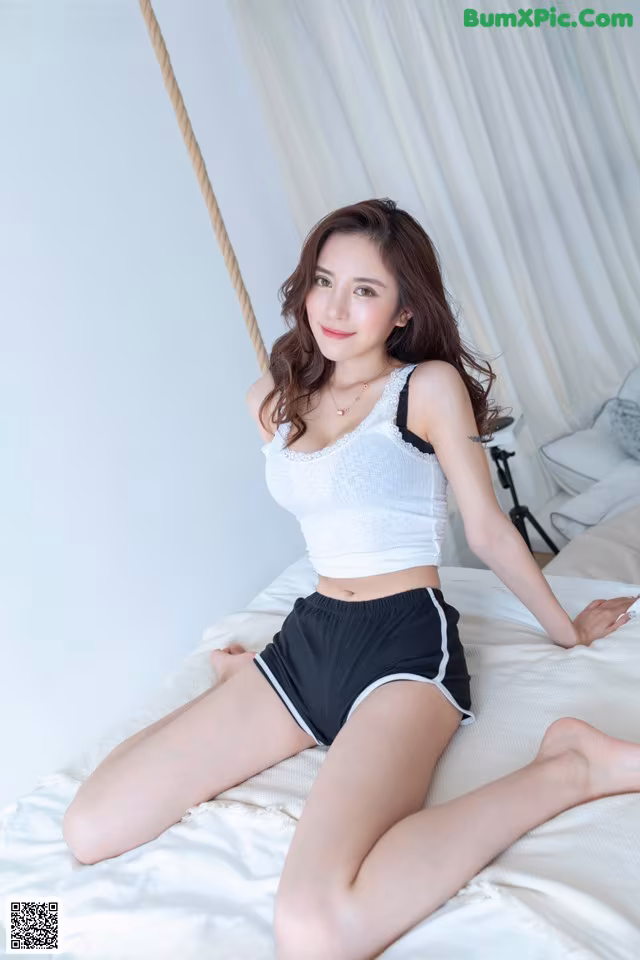 A woman sitting on a bed in a white top and black shorts.