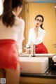 A woman in a red skirt is brushing her teeth in front of a mirror.